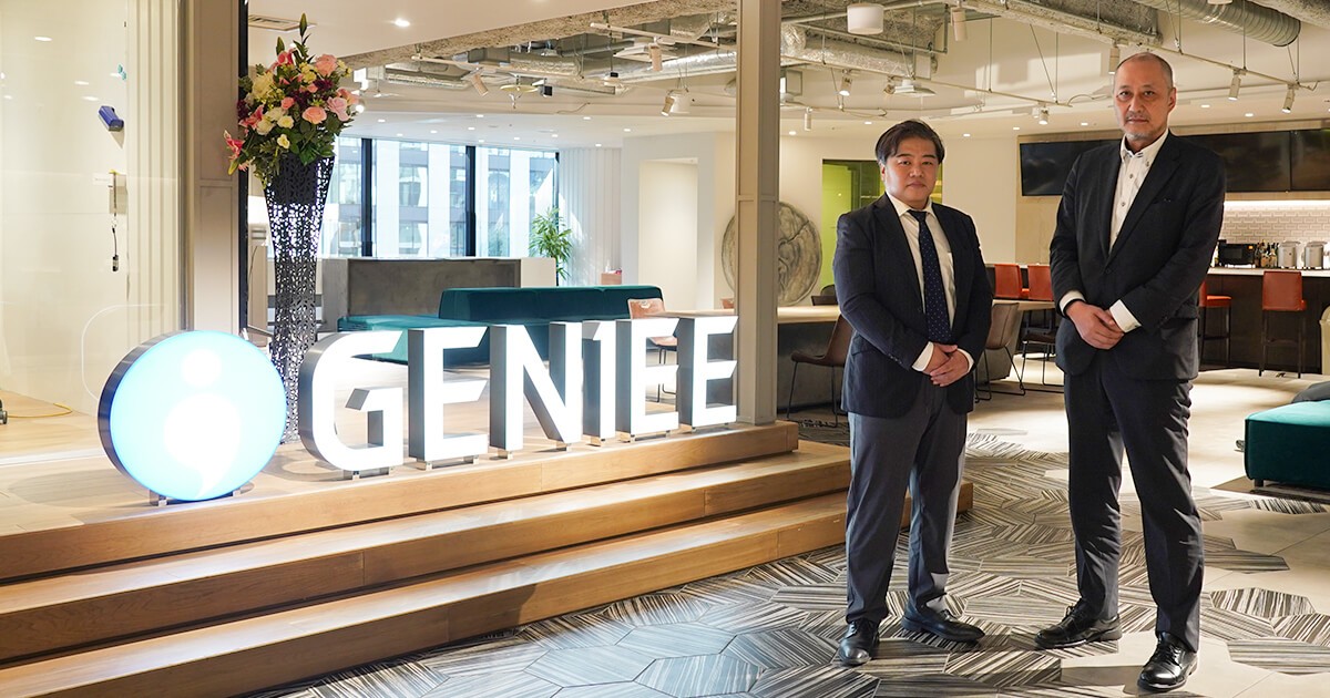 集合写真：株式会社ジーニーのロゴの前で、平井氏と浦が並んでいる様子