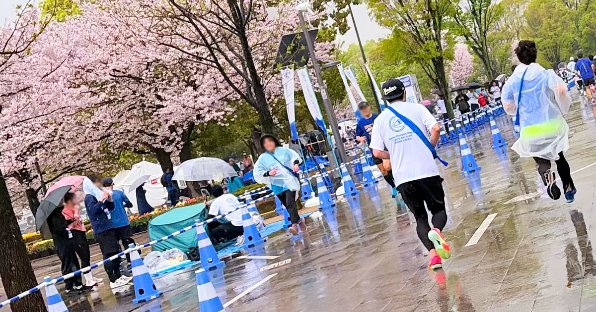 写真:満開の桜を背景に多くの人がコースを走り、沿道では応援をする様子。多くの人が青色を身に着けている。