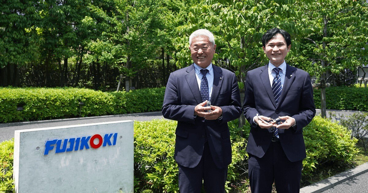 不二工機様玄関の会社ロゴと、草薙様と福井様が並んでいる写真