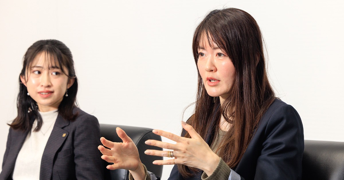 写真：松田 真由氏 / 広報室 コミュニケーションデザイン部（京セラ株式会社）にフォーカスがあたっている。後方から話しを聞いている篠原 花音氏 / 広報室 コミュニケーションデザイン部（京セラ株式会社）。